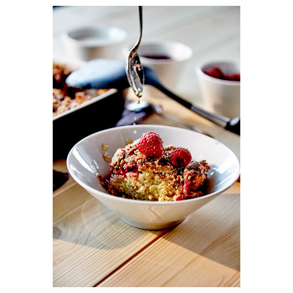 A bowl of fruit crumble on a wooden table. Bowl is white, round, and made of porcelain.