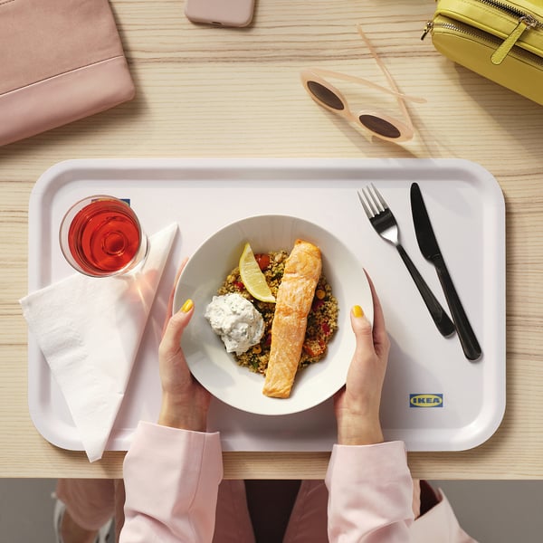 Person holding an IKEA 365+ white bowl with food. The tray holds cutlery, a napkin, and a drink. The setting includes colourful accessories.