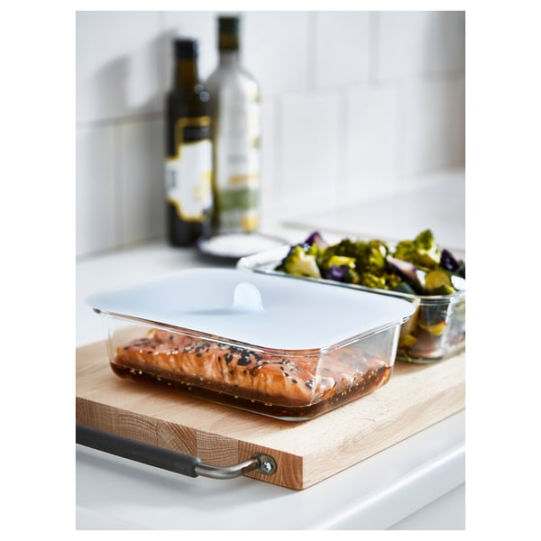 Glass rectangular container with vacuum lid on wood tray, holding cooked fish. Bottles in background.