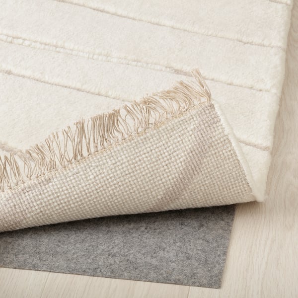 Image shows a section of a cream-colored, textured rug with fringes, placed over a grey rug pad on a light wooden floor.