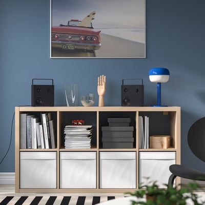 Modern living room with KALLAX wood shelving, books, decor, and vintage car poster against blue wall. Black & white rug beneath.