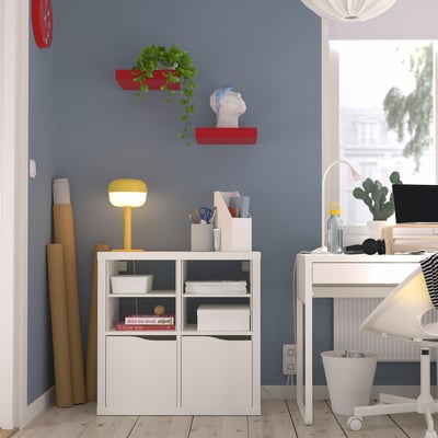 Home office with KALLAX shelf, books, bright lamp, red plant shelves, clock, desk, computer. Blue wall, wooden floor.
