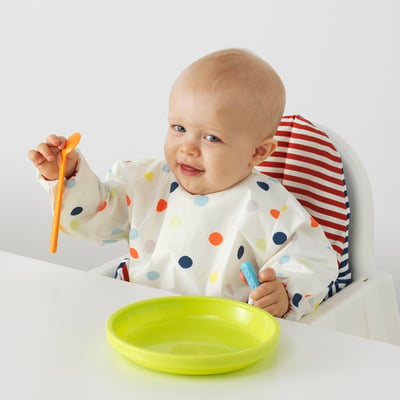 Baby sits in highchair wearing colourful bib with front pocket holding orange spoon and blue baby utensil over green plate.