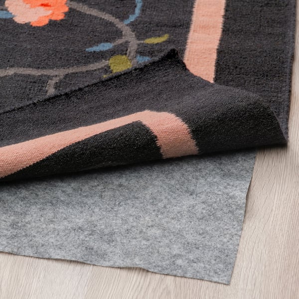 Handwoven wool rug with floral design on dark grey, shown folded with anti-slip underlay on wooden floor.