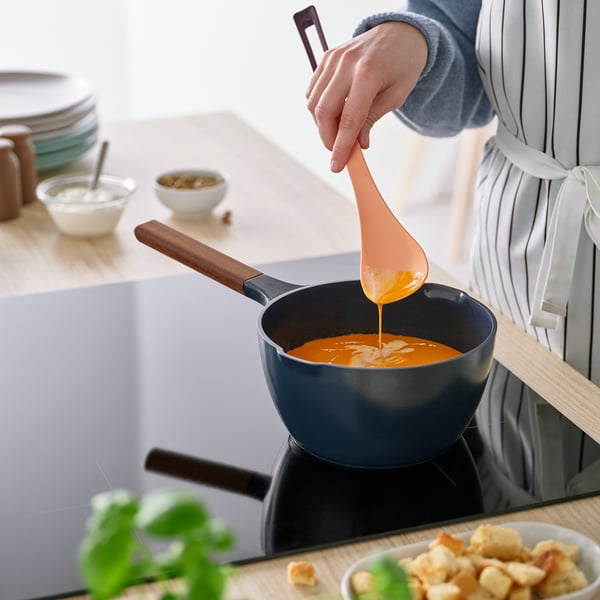 A person stirs orange soup in a blue pan using a colourful spoon with a wooden handle. The spoons narrow design aids precision.