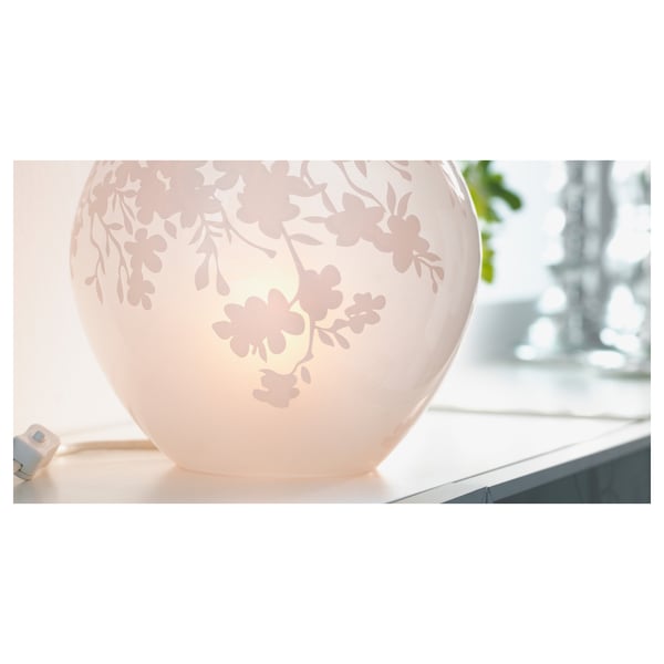 A round, white, table lamp named KNUBBIG with etched cherry blossom design, creating a soft mood light.