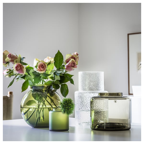 Three glass vases on a white table; left: round, green with flowers; middle: square, clear with candle; right: rectangular, dark green.