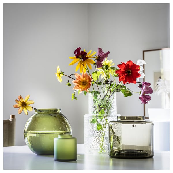 Three vases in green & clear glass, shaped round & square, holding colourful flowers.