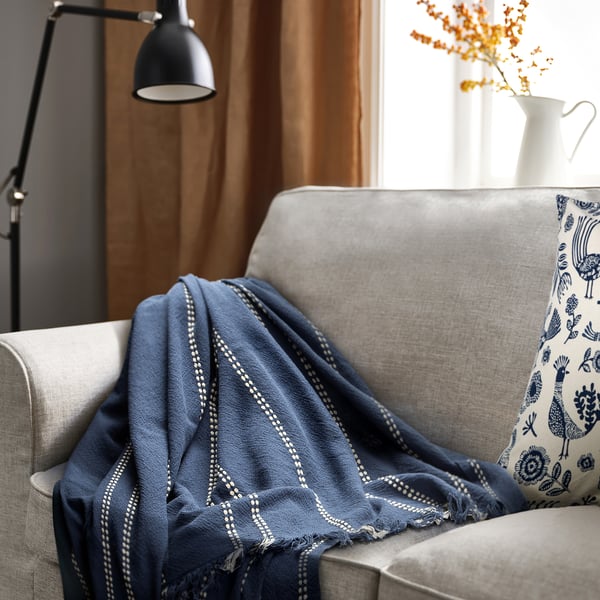 KRUTBRÄNNARE blue throw on grey sofa. Dobby stripe weave, soft cotton, triangular fold, decoration on right armrest.
