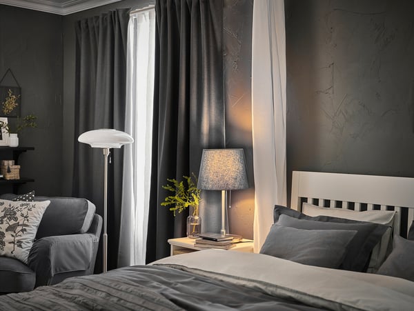 Modern bedroom with KRYSSMAST lamps, grey curtains, striped pillows, cosy bed, and patterned cushion armchair by window.