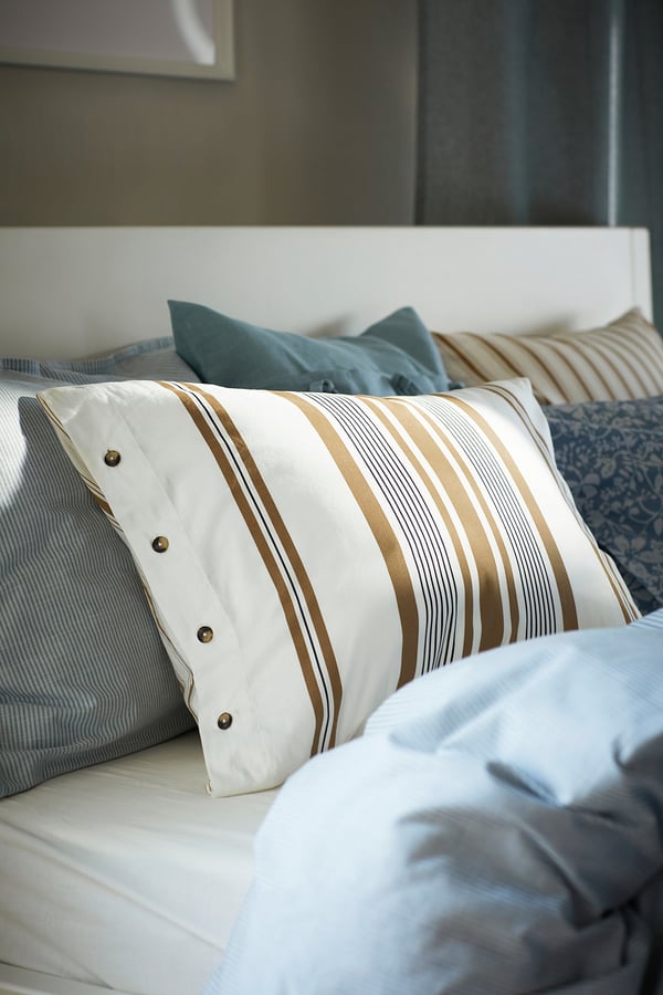 Bed with striped beige and white pillowcases, blue pillows, buttoned design, cosy and inviting.
