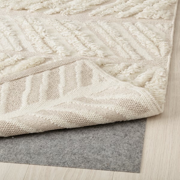 Beige wool rug with geometric pattern and cut furrows, placed on felt rug underlay, showcasing its handcrafted texture.