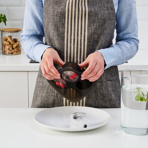 Person holding an ice cube tray with frozen berries over a plate with ice cubes. A glass of water with ice and mint is nearby.