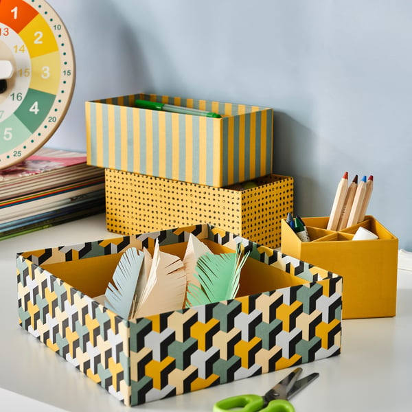 Desk with 4 LYSMASK boxes: yellow, green, black. Feathers, scissors, coloured pencils inside. Stacked, recycled.