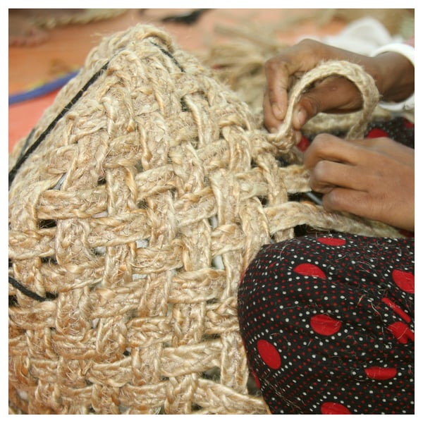 Hands skillfully weave a light brown jute basket on a red-patterned cloth.