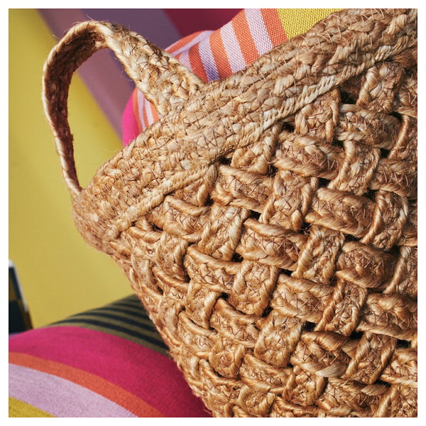 Brown woven jute basket with handles, displayed on colourful fabrics.