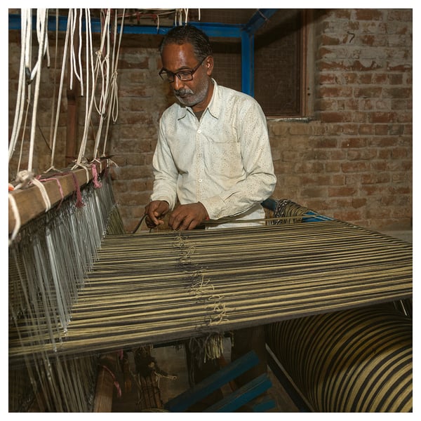 Person weaving striped bedspread on loom in rustic room.