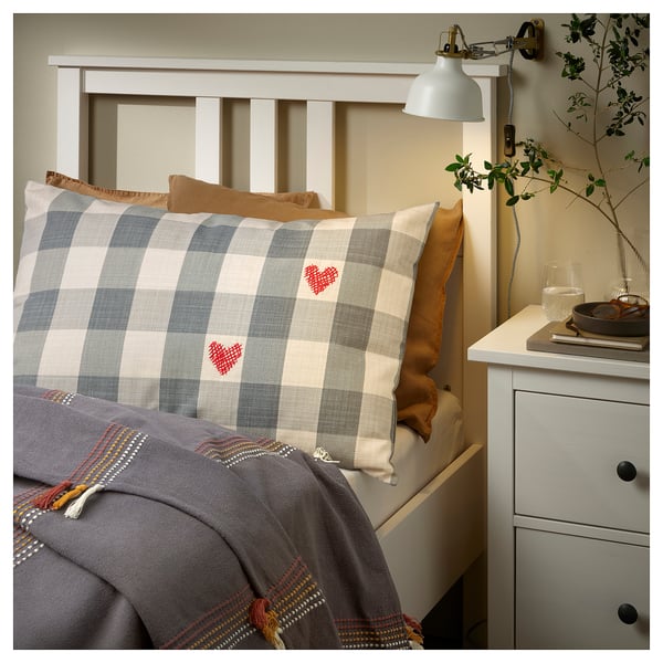 Cosy bedroom with plaid pillow, heart designs, wooden headboard, grey blanket, orange pillow, and bedside table with lamp, vase, glass.