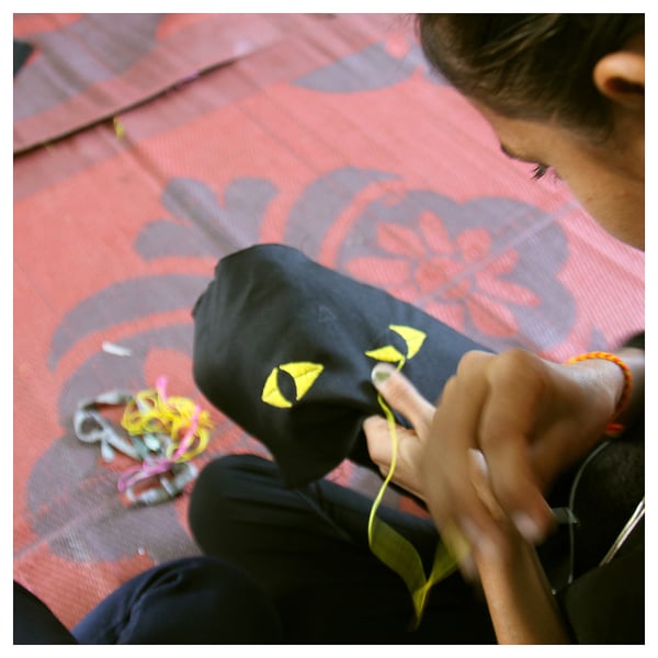A person sews a black cat-shaped pouch on a pink floral cloth. The pouch yellow triangular ears and eyes, and a zip mouth.