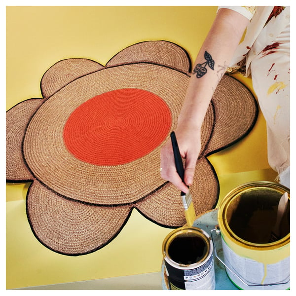 Person painting a large orange circular centre on a woven brown flower-shaped rug on a yellow background.