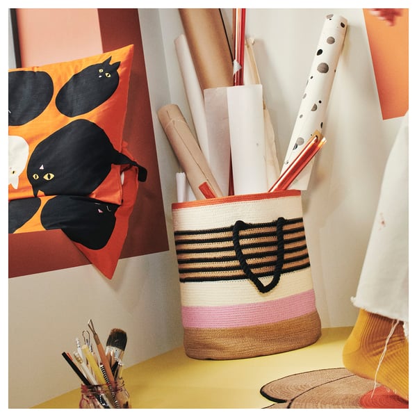 Colourful MÄVINN basket with art supplies on yellow surface, behind black cat fabric.