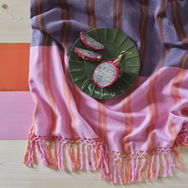 A woven throw blanket with pink and blue stripes and fringe sits on a wooden table. It displays two dragon fruit halves on a green plate.