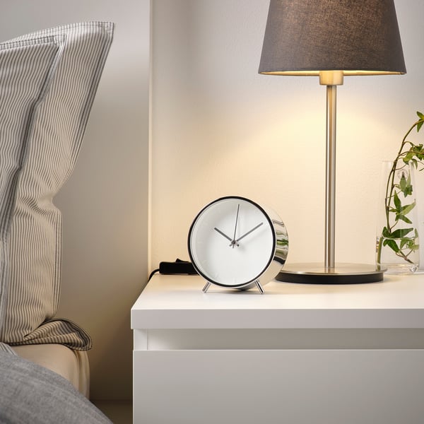 Modern bedside setup with a round alarm clock displaying time, a stylish lamp providing light, and a chair with striped fabric.