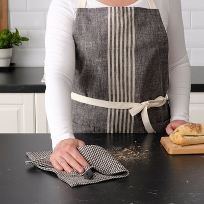 Person in kitchen, wearing a grey striped apron, holding a cloth and cutting bread on a black countertop.