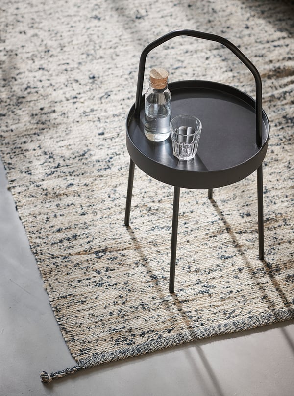 Small black round tray table on a multicoloured braided rug. It holds a glass and bottle.