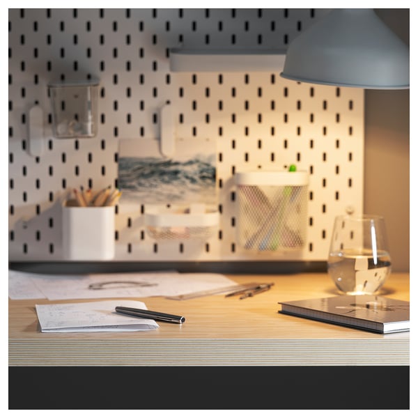Modern home office setup: light wood desk with papers, pen, glass cup, lamp, & storage against white pegboard wall.
