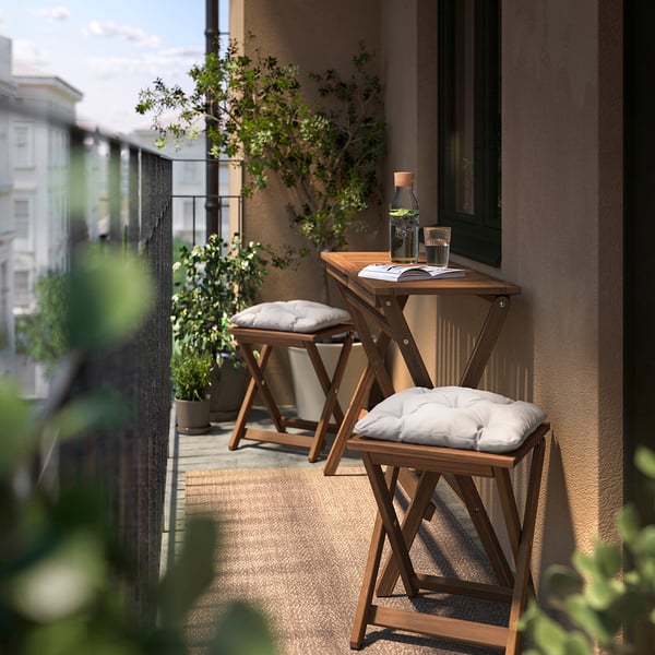 Small balcony with NÄMMARÖ brown foldable wooden table and stools, cushioned in white, placed beside green foliage.