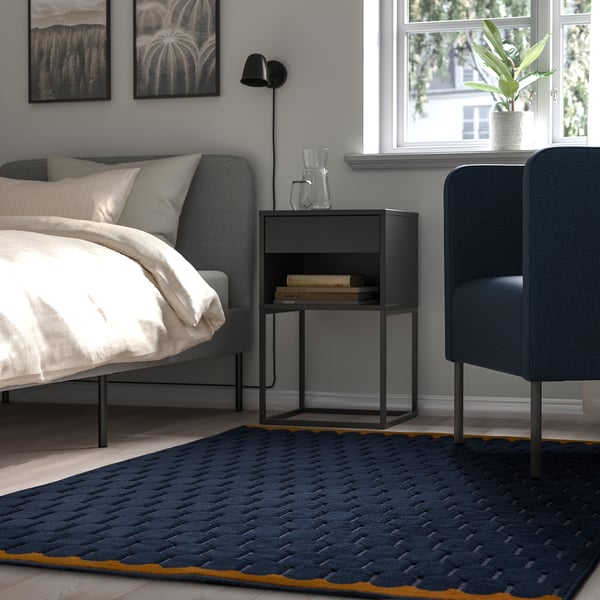 A modern living space with a grey növing rug, black bedside table, and blue upholstered chair, accented with a black wall lamp and framed art.