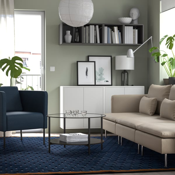 Modern living room with NÖVLING blue rug, grey sofa, blue chair, glass coffee table, white cabinet, books, vases, and plants.
