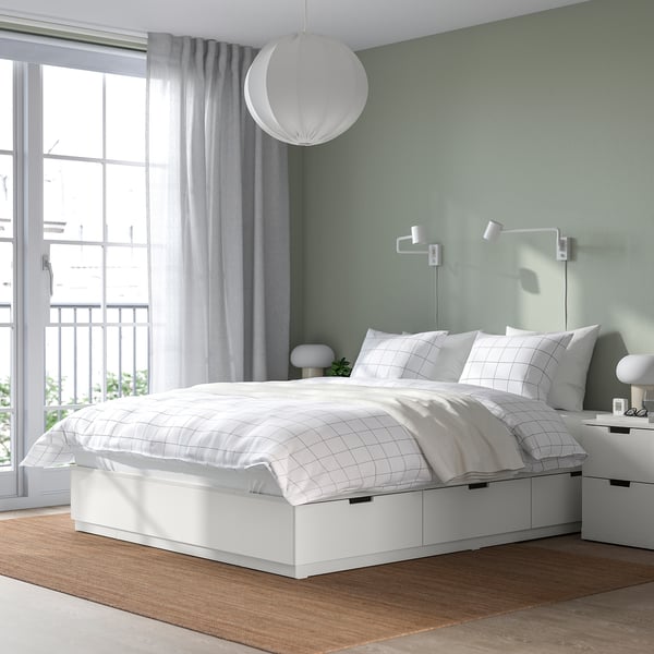 NORDLI bed with 6 drawers in minimalist bedroom. Space-saving, under window with light curtains. Illuminated by ceiling lamp. White bedding.