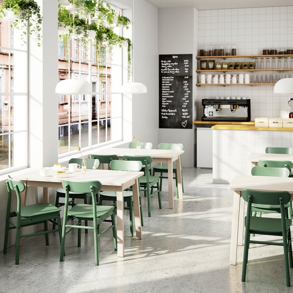 Minimalist café with green chairs, wood tables, large windows, plants, and coffee station.