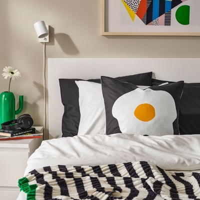 Modern bedroom with white bed, black pillows, green vase, books, and art.