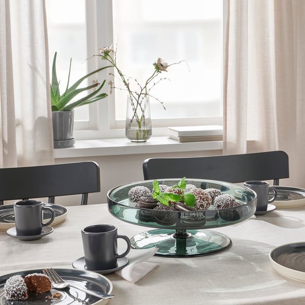 Table set with mugs, plates, cake. Centrepiece is cake stand with desserts. Window plants, white curtains.