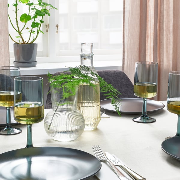 Table set with OMBONAD blue plates, clear textured glass vases holding greenery, and wine glasses on white tablecloth near window.
