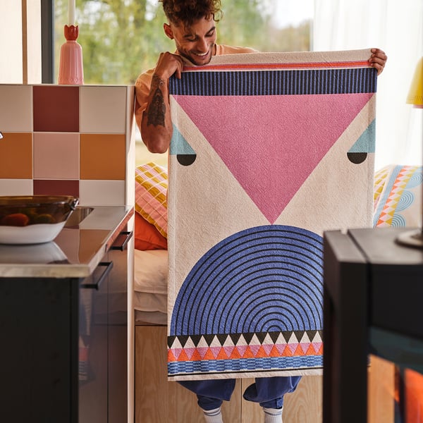 Person holding large, colourful, geometric OMMJÄNGE cotton wall hanging, bigger than nearby kitchen items.