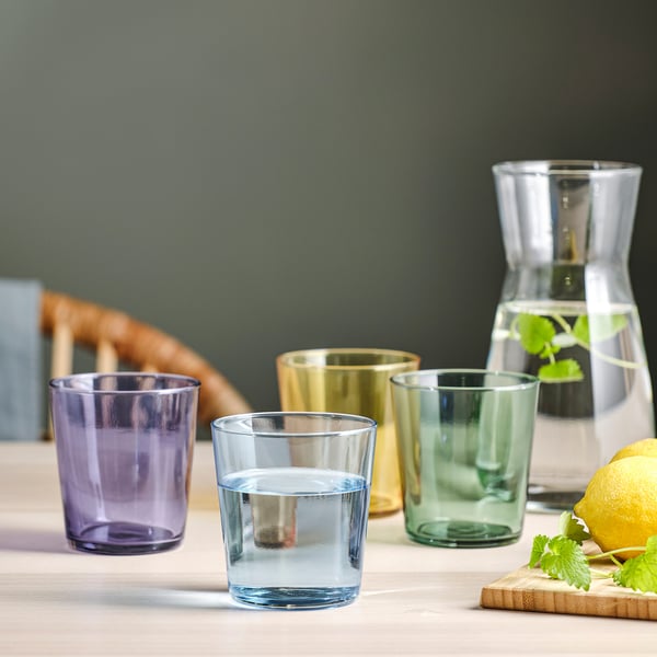 Four coloured glasses and a carafe with water and lemon slices on a table.