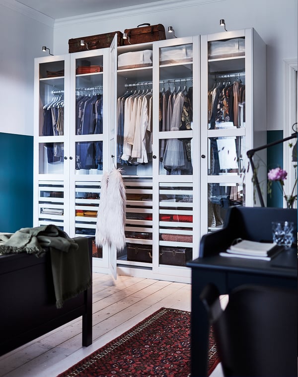 White wardrobe with glass doors, clothes inside, plush coat, wooden floor, red rug, black chair with blanket, desk, hat.