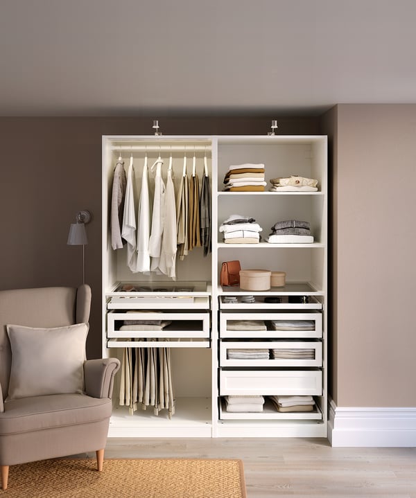Beige PAX wardrobe with clothes, shelves, drawers, glass compartments, and a beige chair by brown walls.
