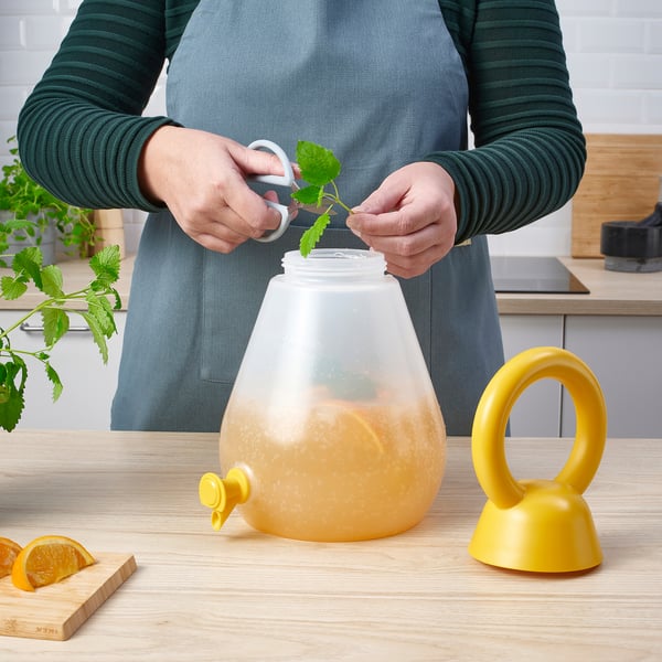 Person places mint leaf into a PILLERSTARR jar with tap, containing a orange drink, in a kitchen setting.