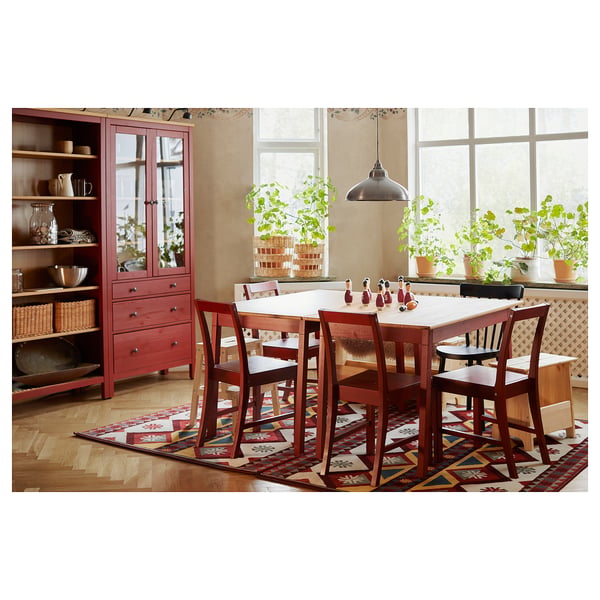 Bright red wooden dining table with four matching chairs. Surrounded by large windows and plants, set on a patterned rug.