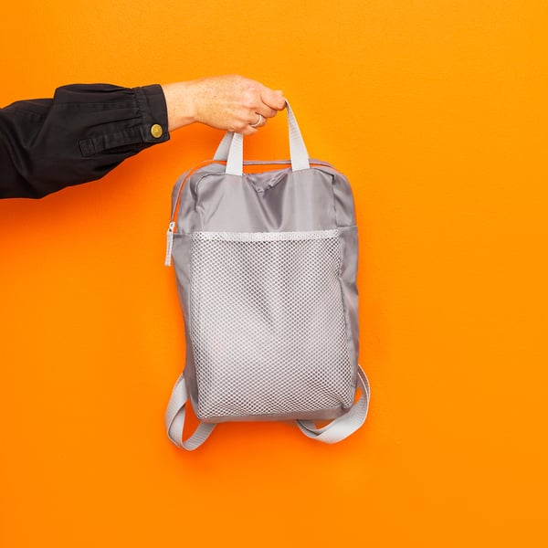 Person holding grey, mesh backpack against bright orange wall, showcasing its design and adjustable straps.