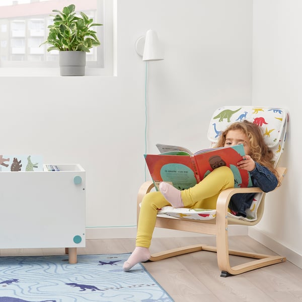 Childs dinosaur-themed POÄNG chair, girl reading, white background.
