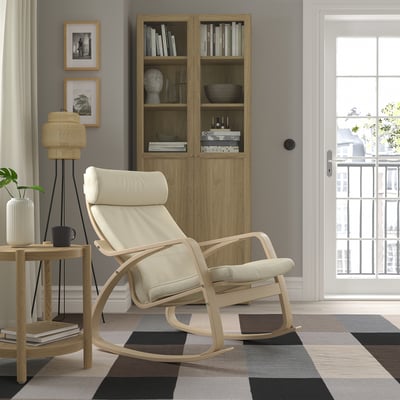 Calm living room with light beige rocking chair POÄNG on colourful chequered rug near wooden bookshelf. Floor lamp left, balcony right.