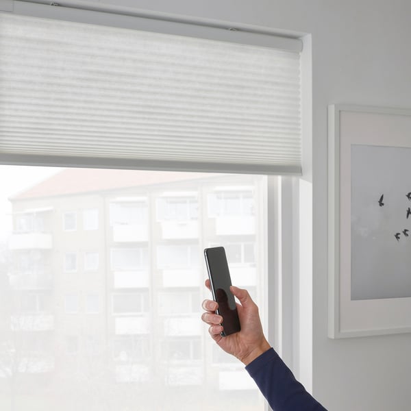 A hand holds a smartphone, controlling smart cellular blinds via an app, demonstrating remote operation.