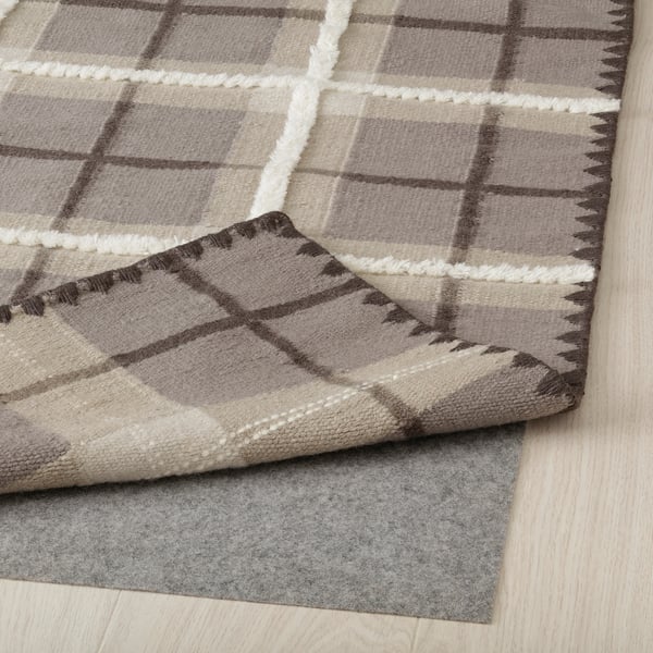 Beige and grey wool rug with tartan pattern, folded over grey felt underlay, showcasing its durable, handwoven design and anti-slip feature.