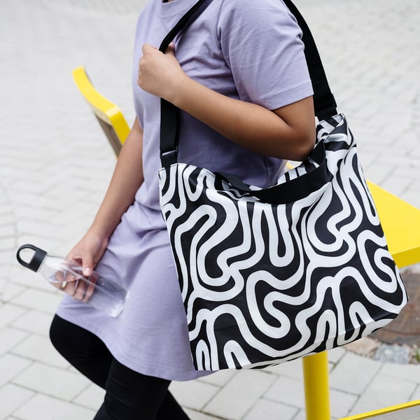 Person carrying black & white RÄCKLA shoulder bag, larger than a water bottle, in abstract pattern, wearing light purple top & black leggings.